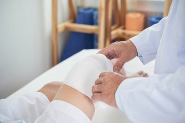 Obraz na płótnie Canvas Anonymous medical practitioner using clean bandage to dress knee of unrecognizable man on blurred background of doctor's office