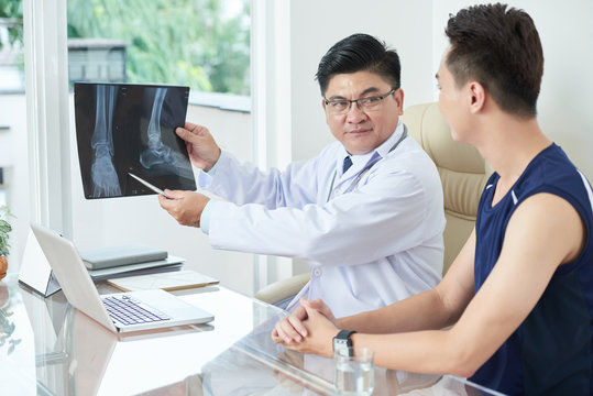 Adult Asian man in medical apparel demonstrating X-ray picture of legs to young guy while sitting at table in doctor's office