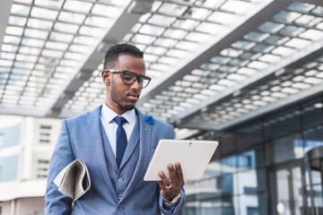 black man businessman in a business suit, expensive watch and glasses with a newspaper and digital tablet goes against the backdrop of a modern city to work