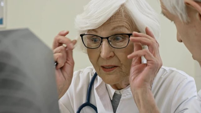 Tilt up of senior female doctor putting on eyeglasses and analyzing x-ray image with elderly male colleague