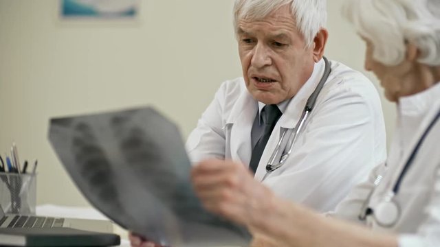 Senior male doctor sitting at desk in clinic and discussing x-ray image with elderly female colleague