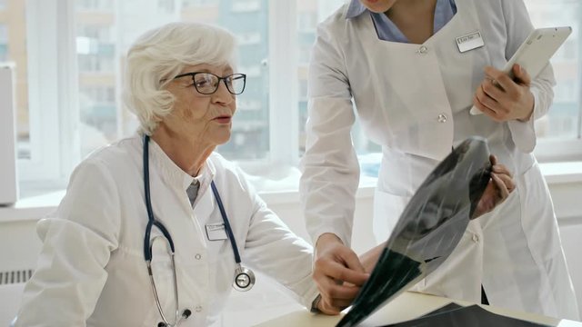 Senior female doctor in lab coat and eyeglasses sitting at desk, looking at x-ray image and discussing diagnosis with female assistant