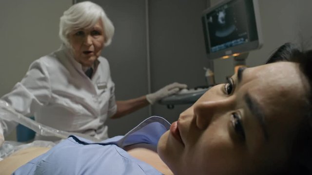 Senior female doctor performing ultrasound exam of abdomen and then telling bad news to Asian female patient