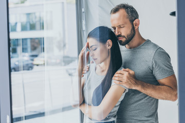 bearded man supporting sad stressed woman with closed eyes standing near window