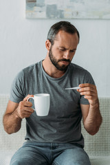 sick bearded middle aged man holding cup and looking at thermometer
