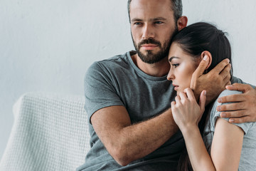bearded man supporting and hugging depressed woman sitting on couch