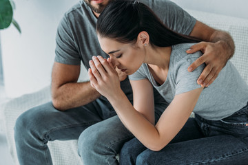 cropped shot of man supporting upset girlfriend while sitting together on couch