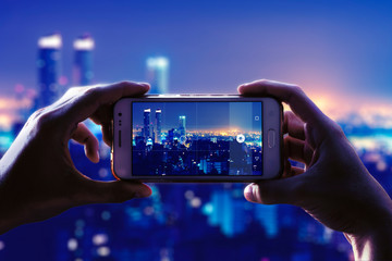 Photo camera of a smartphone. View through the screen at the time a young woman takes a photo of a city at night. Cityscape.