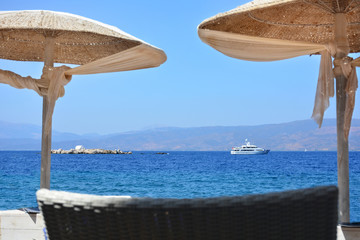 Small island with wedding chapel and ship in the sea visible through beach umbrellas from the shore in Hydra island, Greece