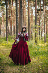 Fototapeta premium A beautiful girl with black long hair in a medieval, red dress with a crown on her head walks in the forest. Fairy-tale princess. Fantasy