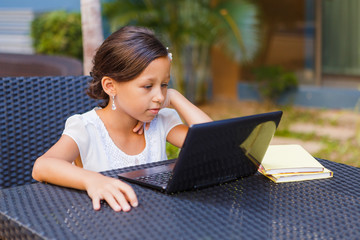 A beautiful, elegant girl of 8 years sitting with a laptop and books outdoors, studying at an online school
