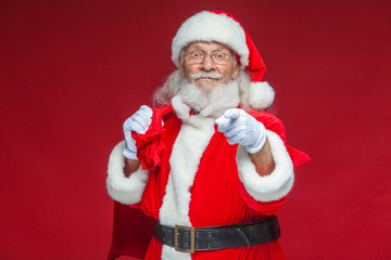 Christmas. Smiling Santa Claus in white gloves with a bag of gifts behind him points his index finger into the camera. Isolated on red background.