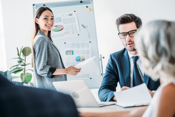 smiling asian businesswoman standing near flipchart during meeting in office