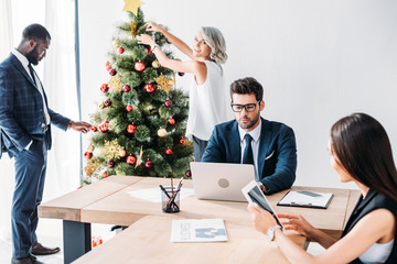 Young multicultural businesspeople working and decorating Christmas tree in office