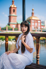 Beautiful girl with black long hair in a white dress on a bridge, walk near a pond. An orthodox woman