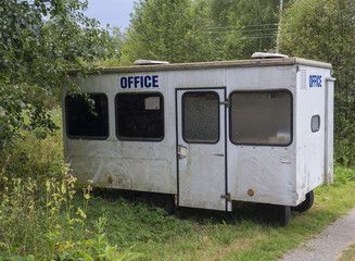Small white trailer house with blue sign office, abandoned caravan in rural landscape near footpath, green grass and trees