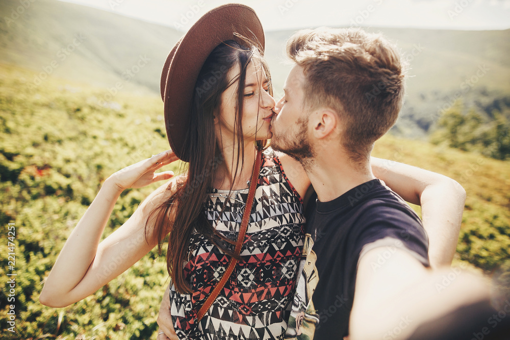 Wall mural stylish hipster couple taking selfie and kissing on top of mountains in sunny summer day. beautiful 