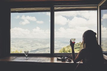 stylish girl traveler sitting at window with view on mountains and sky. happy hipster woman holding drink, looking at sky and relaxing. summer vacation concept. travel and wanderlust