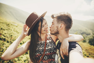 stylish hipster couple taking selfie and kissing on top of mountains in sunny summer day. beautiful man and happy woman in hat embracing on honeymoon. travel together