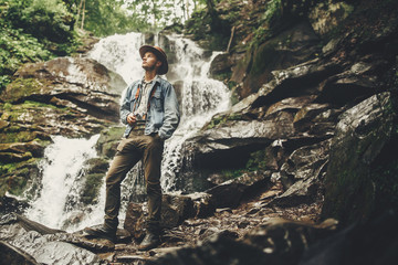 stylish hipster man in hat with photo camera, standing at waterfall in forest in mountains. traveler guy exploring woods. travel and wanderlust concept. atmospheric moment