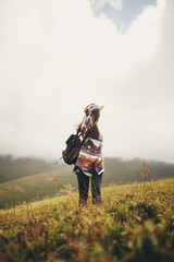 travel and wanderlust concept. stylish traveler hipster girl holding hat, with backpack and windy hair, walking in mountains in clouds. summer vacation. space for text. amazing atmospheric moment