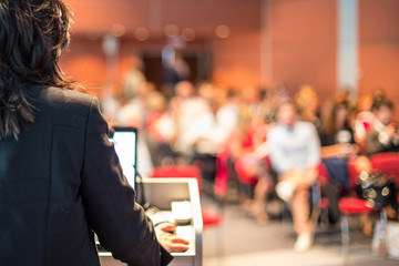 Female speaker giving a talk on corporate business conference. Unrecognizable people in audience at...