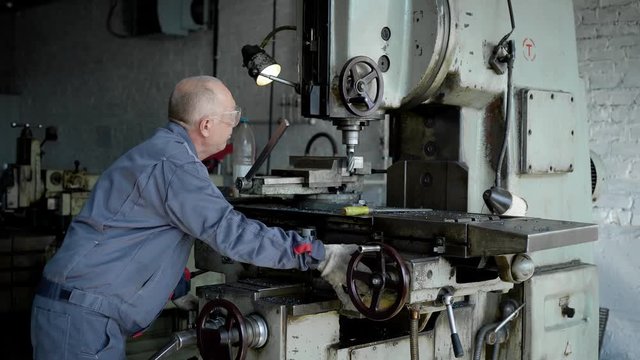 An elderly man in production. A worker on a machine drills a metal part.