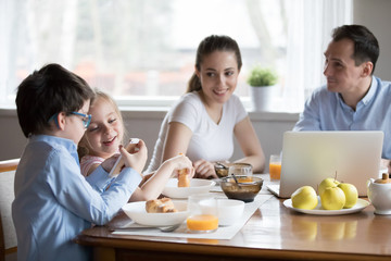 Cute excited kids playing on smartphone while having homemade croissants in morning, happy parents watch children talk busy with cell enjoying tasty breakfast at home, spending time together