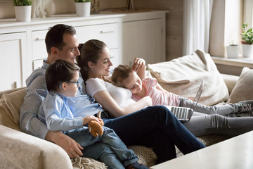 Happy parents hug kids lying on cozy sofa watching cartoons on laptop, young family spend time with...