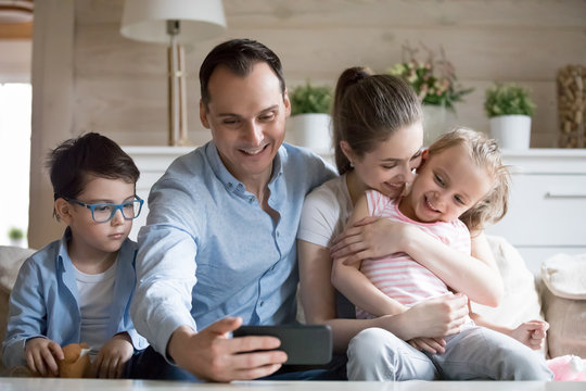 Happy family of four sitting on couch together posing for selfie in living room, smiling father hold smartphone taking picture of parents with kids, mom dad and children look at camera making photo
