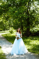 Portrait of beautiful brunette bride with curly hair in Park on green foliage background. Girl in a lush blue wedding dress with a bouquet of flowers.