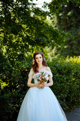 Portrait of beautiful brunette bride with curly hair in Park on green foliage background. Girl in a lush blue wedding dress with a bouquet of flowers.