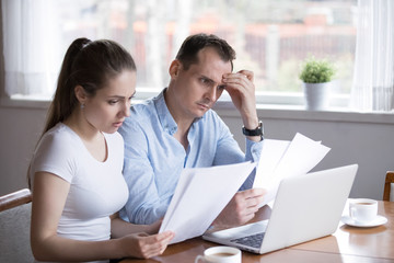 Serious husband and wife reading paper documents analyzing house utility bills, stressed couple get...