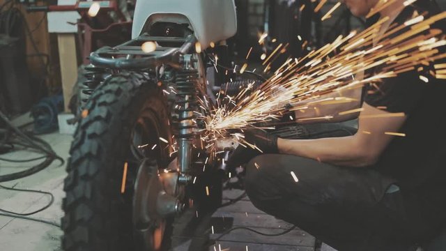 Man works circular saw. Flies of spark from hot metal. Handsome guy working with custom motorcycle in the garage. Close-up of hand and electric saws metal hand tool