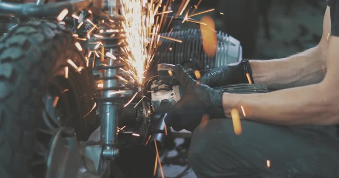 Man works circular saw. Flies of spark from hot metal. Handsome guy working with custom motorcycle in the garage. Close-up of hand and electric saws metal hand tool