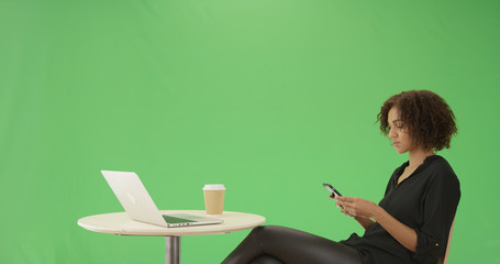 A young black woman sits at a cafe table on her phone on green screen