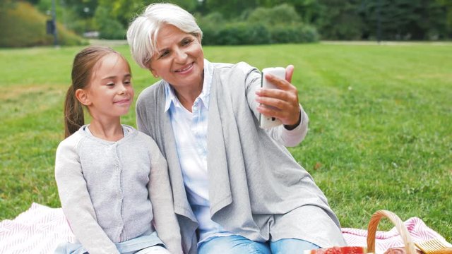 family, leisure and technology concept - happy grandmother and granddaughter having picnic and taking selfie by smartphone at summer park