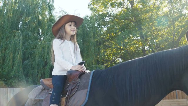 Pretty little girl in hat riding a black mare on the arena. 4K