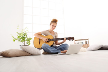 Beautiful smiling young woman playing guitar with computer, learn to play with an online course, sitting on the floor in casual clothes at home