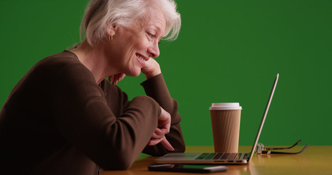 Side profile of happy old woman scrolling on laptop with coffee on green screen