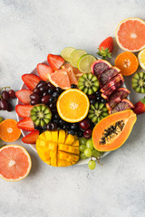 Raw fruits berries platter, mango, oranges, kiwi strawberries, blueberries grapefruit grapes, apples on the white plate, on the off white table, top view, vertical, copy space