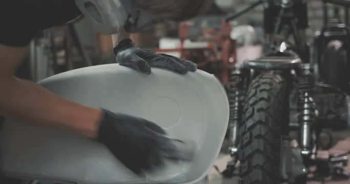 Guy polishing metal gas tank with sandpaper. Auto mechanic assembles custom motorcycle in his workshop. Guy create an exclusive motobike cafe racer in the garage.