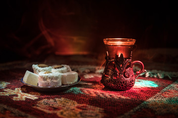 Arabian tea in glass with eastern snacks on a carpet on dark background with lights and smoke. Eastern tea concept. Empty space.