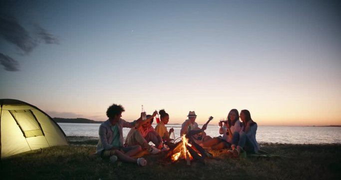 Group of six old friends having a summer beach party near bonfire, talking, smiling, drinking beer, wine, toasting and cheering having fun 4k