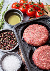 Plastic tray with raw minced homemade beef burgers with spices and herbs. Top view. On top of  kitchen table background with tomatoes salt and pepper.