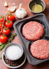Plastic tray with raw minced homemade beef burgers with spices and herbs. Top view.On rusty kitchen table background with tomatoes salt and pepper.
