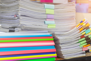 Close up pile of unfinished paperwork on teacher's desk waiting to be managed. Stack of homework assignment separated by colorful paper clips and plastic binding bars. Education and business concept.