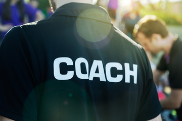 Sport coach in black shirt with white Coach text on the back standing outdoor at a school field, with morning lens flare - obrazy, fototapety, plakaty