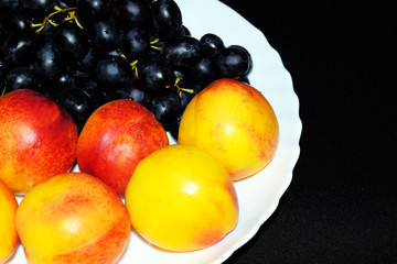 Peaches, black seedless grapes and nectarines. Orange juice and fruit for Breakfast. Yellow tropical fruit on a white plate. A glass of juice on a black background. Vegetarian and raw food menu. Diet