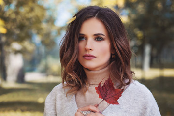 Autumn beauty. Cute woman outdoors. Young female model with healthy hair, makeup and fall leaves in hand walking in autumn park